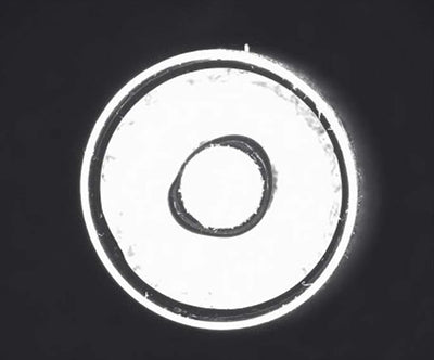 Source capture of a cylindrical battery part, end-on, under a red dome light: a bright face with an irregular dark ring around the central opening and a thin dark ring near the rim. Complete capture inside the brochure frame, not retouched.