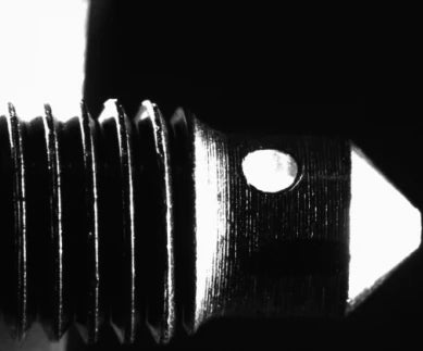 Source monochrome capture of a threaded pin with a cross hole near its tip under a high-intensity spot light: bright hole, tip and thread crests. Complete capture, not retouched.