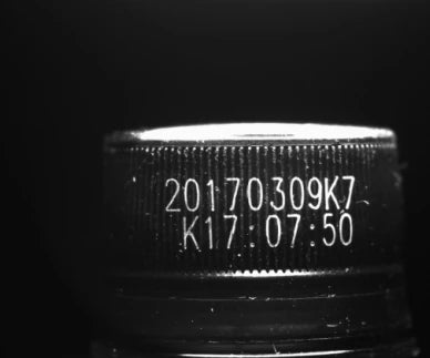 Source capture of the ribbed side of a dark bottle cap under an 850 nm infrared ring light: a two-line date and time code in bright characters. Complete capture, not retouched.