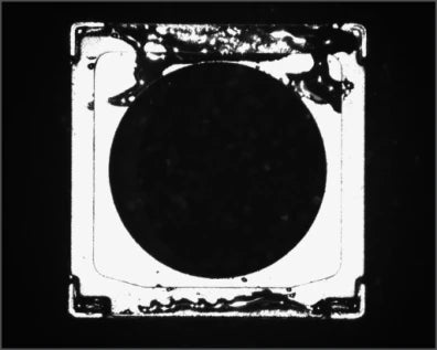 Source capture of a square camera-module frame: the bright flat face surrounds a dark circular opening, with irregular dark glue deposits along the upper and lower edges. Complete capture, not retouched.