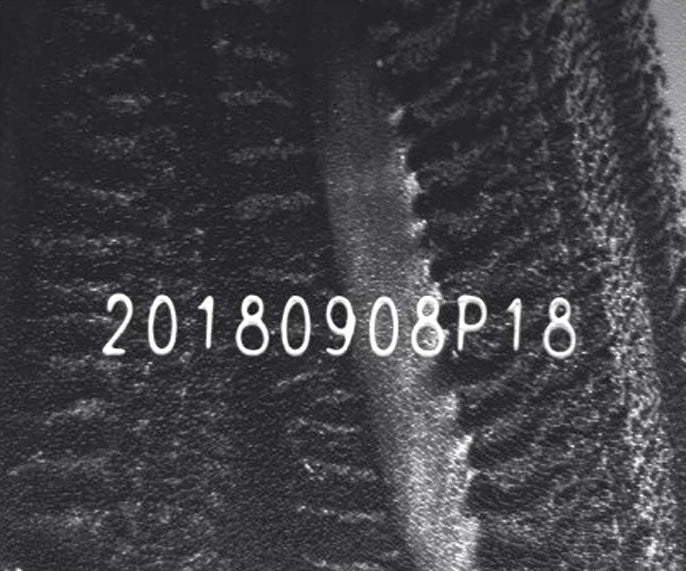 Source capture of corrugated board under a blue bar light: a light-coloured dot-printed date and batch code across the fibre texture and a raised ridge. Complete capture inside the brochure frame, not retouched.