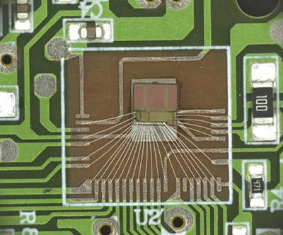 Source colour capture of a bare die on a green circuit board under a white coaxial light: fine wires fan out from the die to the board pads, with small chip resistors nearby. Complete capture inside the brochure frame, not retouched.