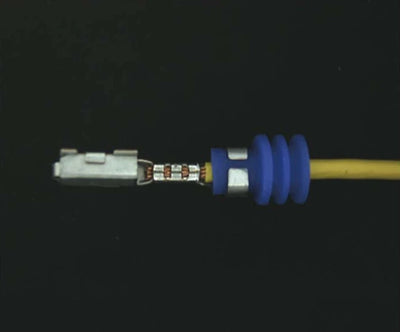 Source colour capture of a crimped wire terminal under a white polarized ring light: metal terminal and crimps, blue seal and yellow wire on a dark background. Complete capture inside the brochure frame, not retouched.