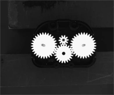 Source capture of an open gearbox under a blue shadowless ring light: four white gears of different sizes in a black housing. Complete capture, not retouched.