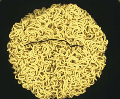 Source colour capture of a round instant-noodle cake under a white dome light: a thin dark fibre lies across the yellow noodles. Complete capture inside the brochure frame, not retouched.