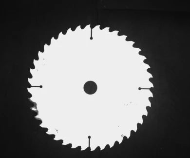 Source monochrome capture of a circular saw blade under a red panel light: bright face, dark bore, four radial slots and a few dark marks on the face. Complete capture, not retouched.